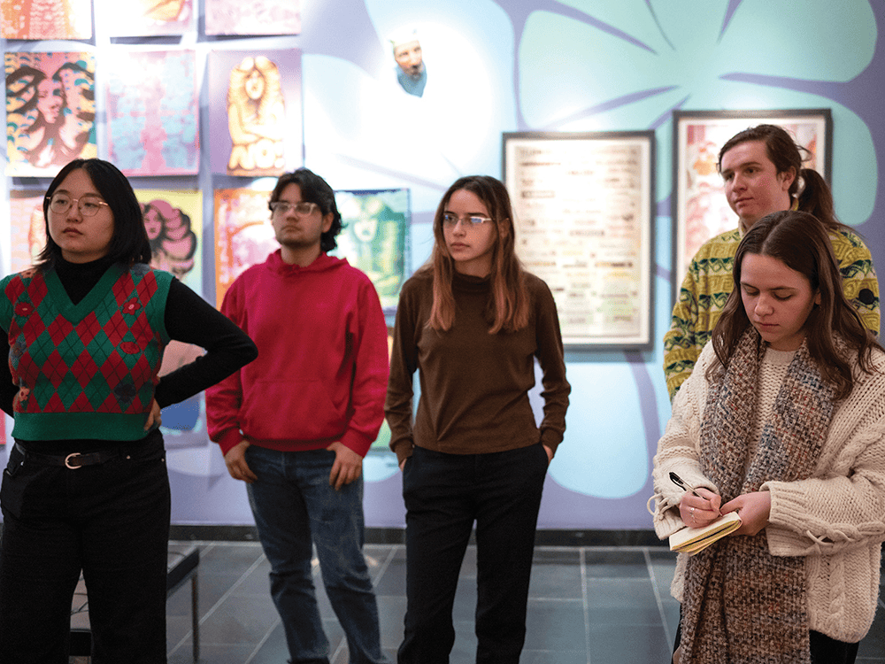 A photo of five Public Humanities student interns standing in the Institute for the Humanities Gallery space in front of Nicole Marroquin's With Care exhibition.
