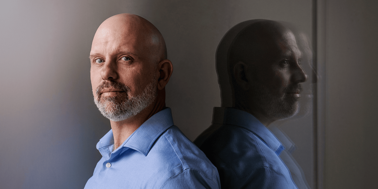 Sociology faculty member Matthew Sullivan poses in a light blue dress shirt, with his reflection visible to the right of his head. His expression is serious. 