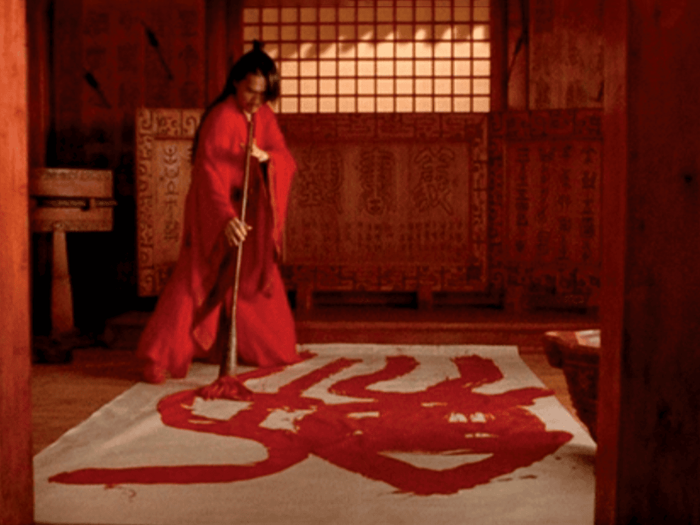 A character in the movie Hero wears a long red robe and paints the character character “jian,” which means “sword,” in red paint on the floor. The character is surrounded by walls that glow with red light.