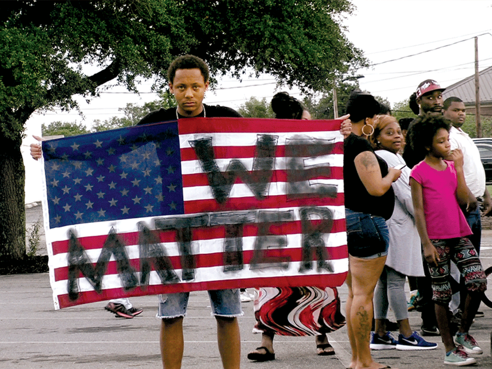 A movie still from a Brave New Films production in which a person of color holds an American flag painted with the words "We Matter."