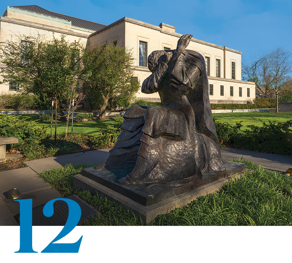 A sculpture of a human figure, seated and with its head in its arms, is pictured outside a building; a stylized number 12 is also pictured.