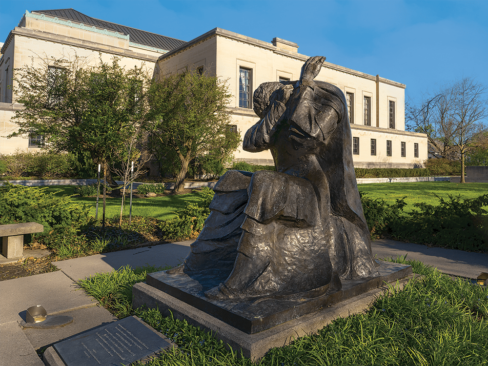 A sculpture of a human figure, seated and with its head in its arms, is pictured outside a building.