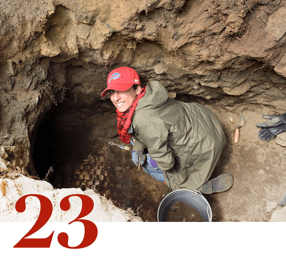 A person looks up from a hole in the ground where she is surrounded by archaeological tools. A stylized numeral 23 is also pictured.