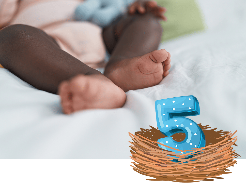 A baby rests on a blanket with their tiny feet in focus. A polka-dotted numeral 5 sits in a nest in the foreground.