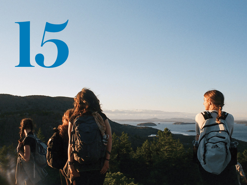 Four people are pictured from behind, all wearing backpacks, overlooking a mountainside and a lake. A stylized numeral 15 is also pictured.