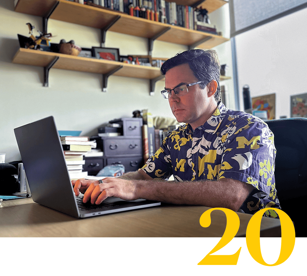A faculty member wearing glasses sits at a desk, typing on a laptop. He wears a Hawaiian-style shirt with Michigan Block Ms on it. A a stylized numeral 20 is also pictured.