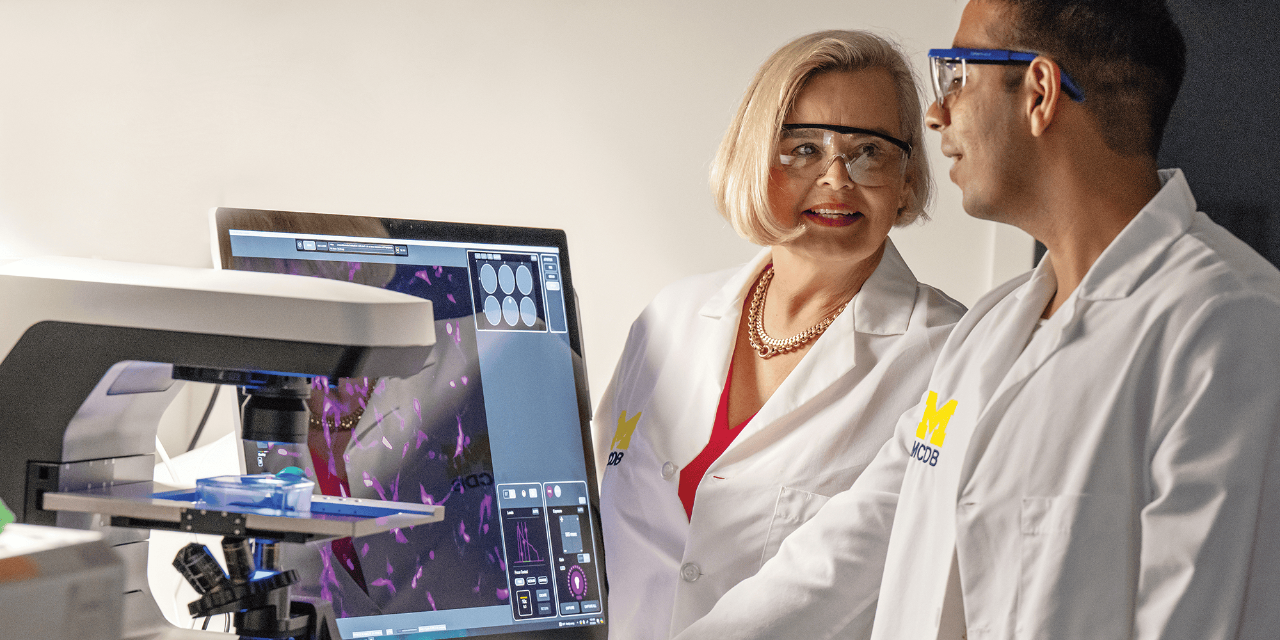 Two people wearing safety goggles and white lab coats, talking with each other, standing next to a microscope and a computer monitor showing an image of cells.