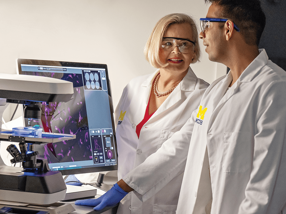 Two people wearing safety goggles and white lab coats, talking with each other, standing next to a microscope and a computer monitor showing an image of cells.