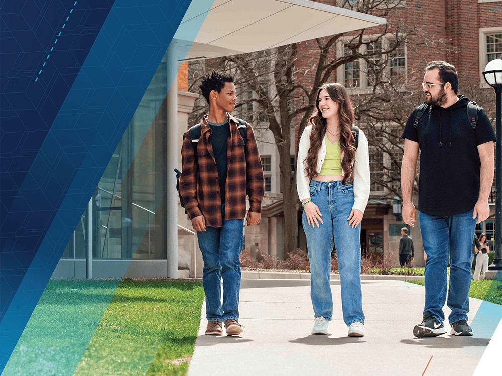 Three students walk down a sidewalk with the LSA Building and Michigan Union in the background. They are looking at each other and appear to be in conversation.