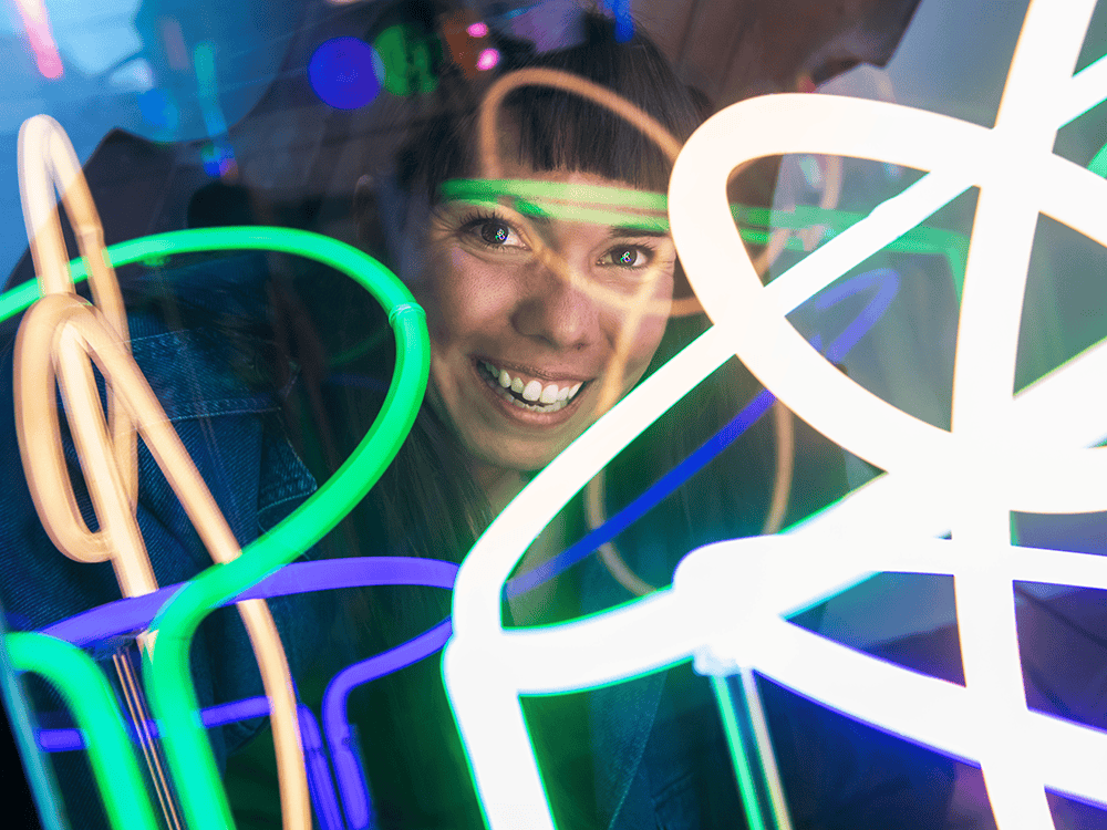 A photograph of Julianna Loera-Wiggins’s smiling face. She has dark hair and wears a denim jacket. Neon lights flash over her face. 