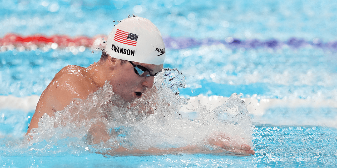 Charlie Swanson swims breaststroke in a pool, his mouth open as he takes a breath. He wears goggles and a white swim cap with an American flag.