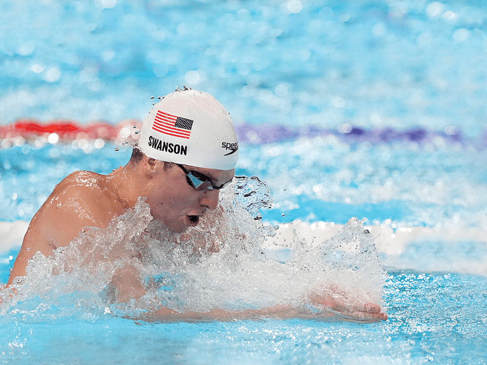 Charlie Swanson swims breaststroke in a pool, his mouth open as he takes a breath. He wears goggles and a white swim cap with an American flag.