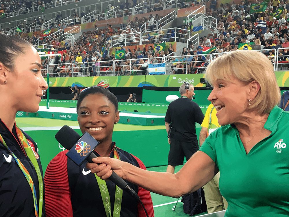 Sports broadcaster Andrea Joyce in profile, holding a microphone to gymnast Aly Raisman, with gymnast Simone Biles at her side. Joyce wears a green shirt that says “Rio.”
