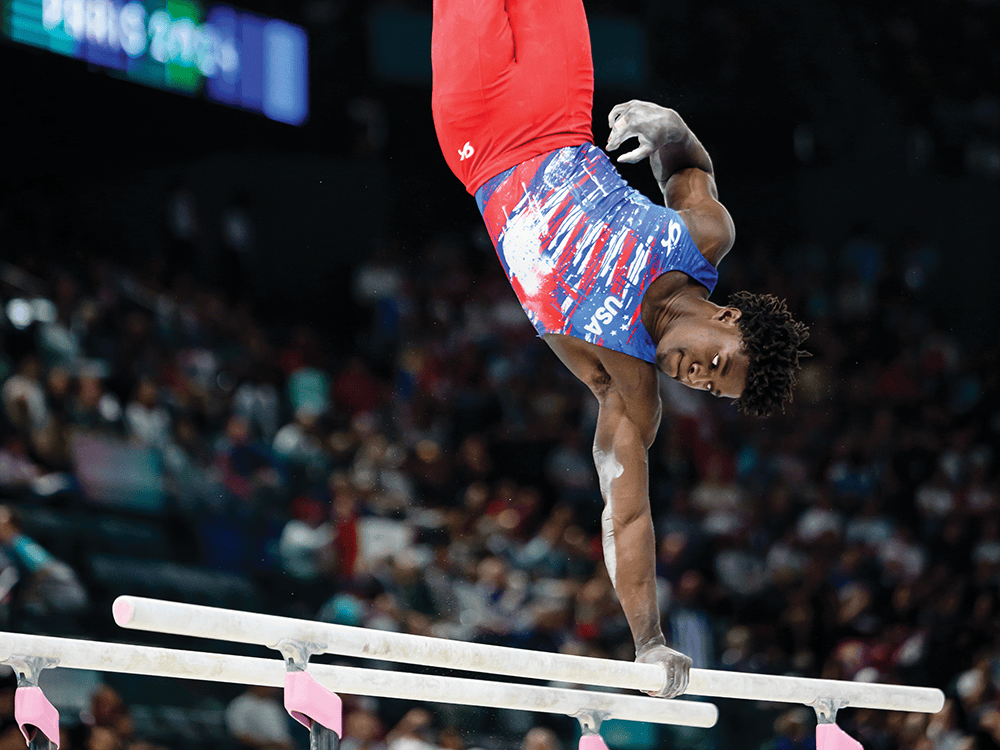Gymnast Frederick Richard holds himself up with one arm on the parallel bars, his eyes gazing down toward the bars. He wears the Team USA uniform with a blue shirt and red pants.