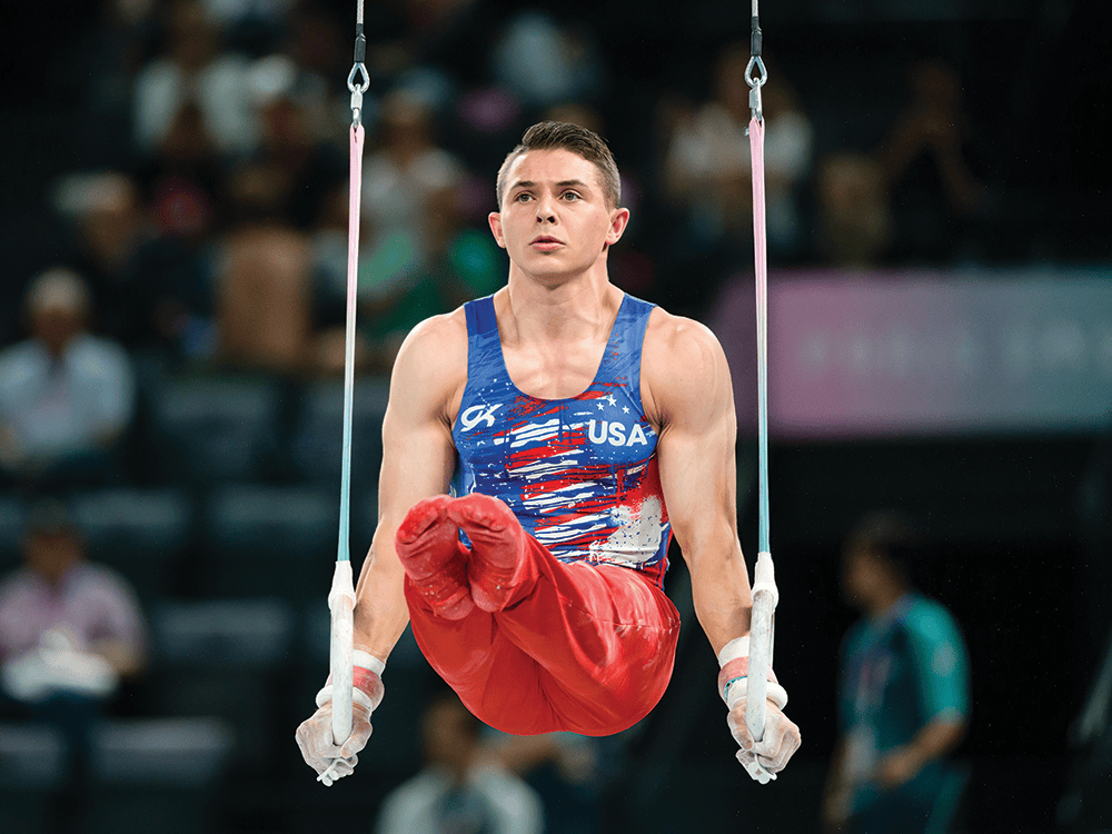 Paul Juda competes on the gymnastics rings event, holding his arms straight down and his legs straight out in front of him. He wears the Team USA uniform with a blue shirt and red pants.