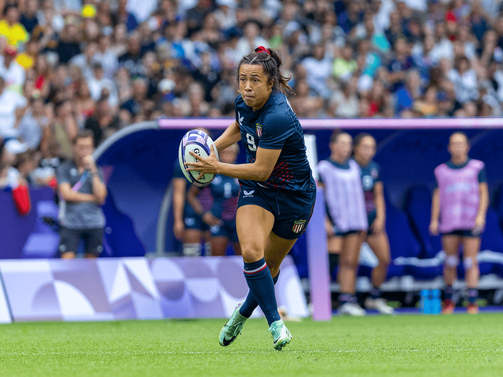 Rugby player Alena Olsen runs with the ball during a match. She wears a blue USA uniform and a red bow in her hair. A crowd and other players are blurry in the background.