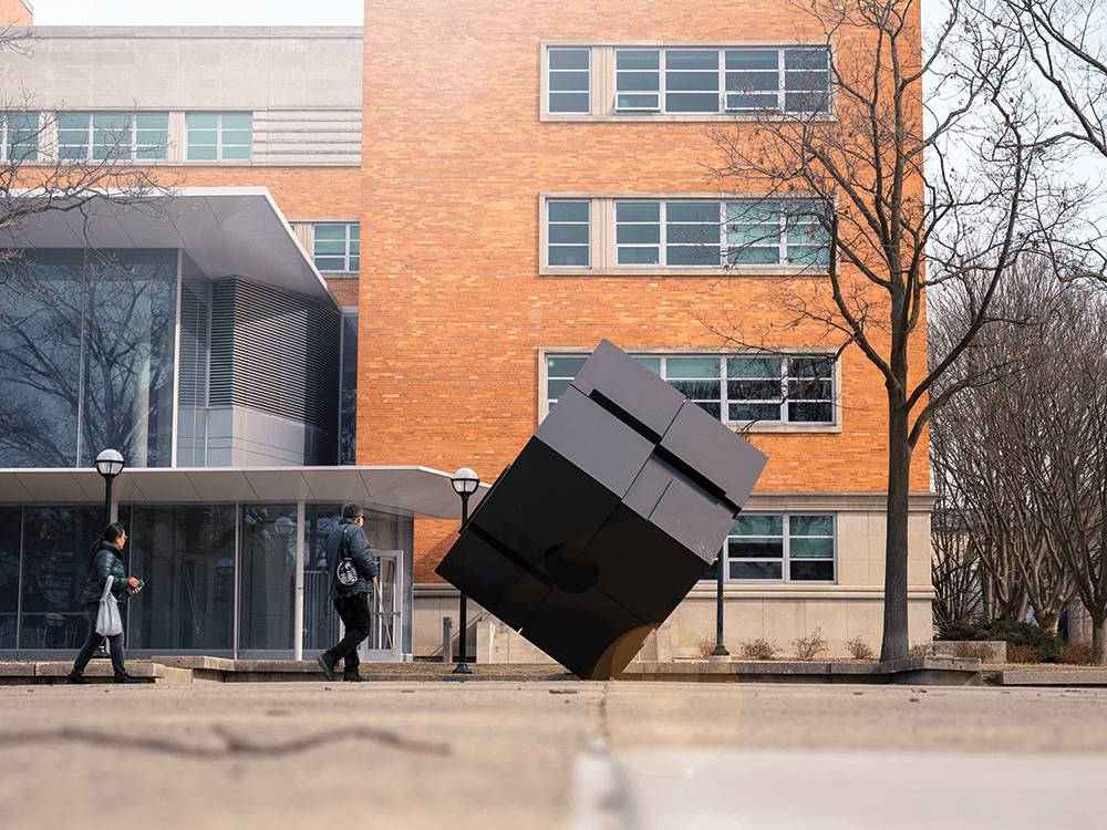 A photo of the exterior of the LSA Building, with The Cube sculpture in the foreground and two people walking nearby.