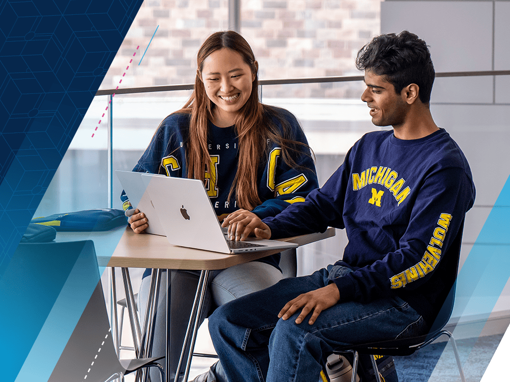 Two students sit at a table in front of a glass wall. They are smiling and looking at the screens of their laptops. Both wear blue shirts with “Michigan” in yellow type.