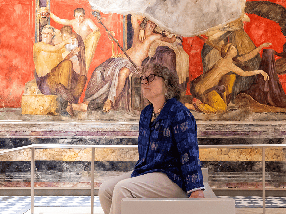 A person in a blue shirt and glasses sits in the center of a room, surrounded by walls painted in the style of an ancient fresco.