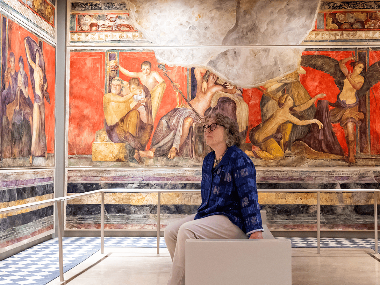 A person in a blue shirt and glasses sits in the center of a room, surrounded by walls painted in the style of an ancient fresco.