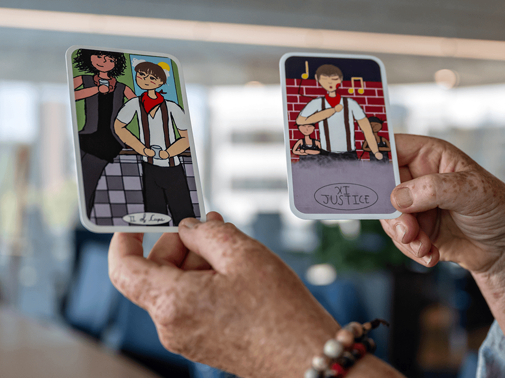 Two hands hold up two tarot cards. One card reads“Two of Cups” and has two figures. The other reads “Justice” and has a singing figure.