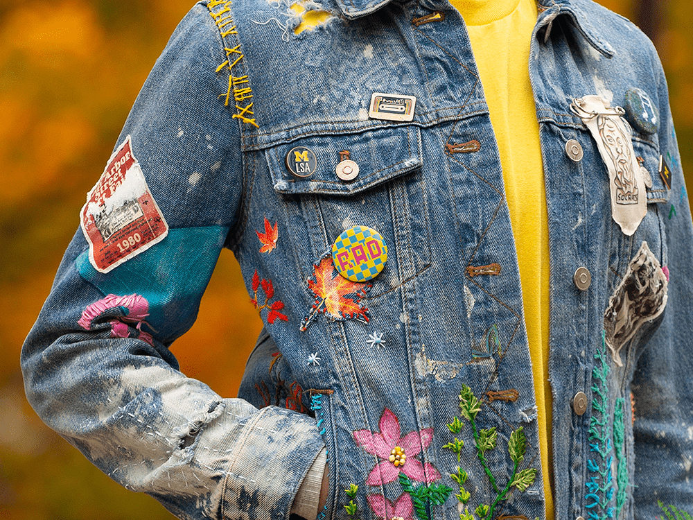 A photo of a person wearing a denim jacket. The jacket includes embroidered flowers, a variety of pins and buttons, paint, a handmade fabric piece with a drawing of Socrates, and faded spots that suggest many years of wear. 
