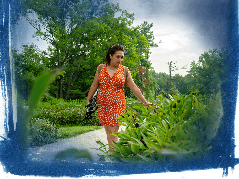 A person in an orange dress walks through an outdoor space with green foliage.