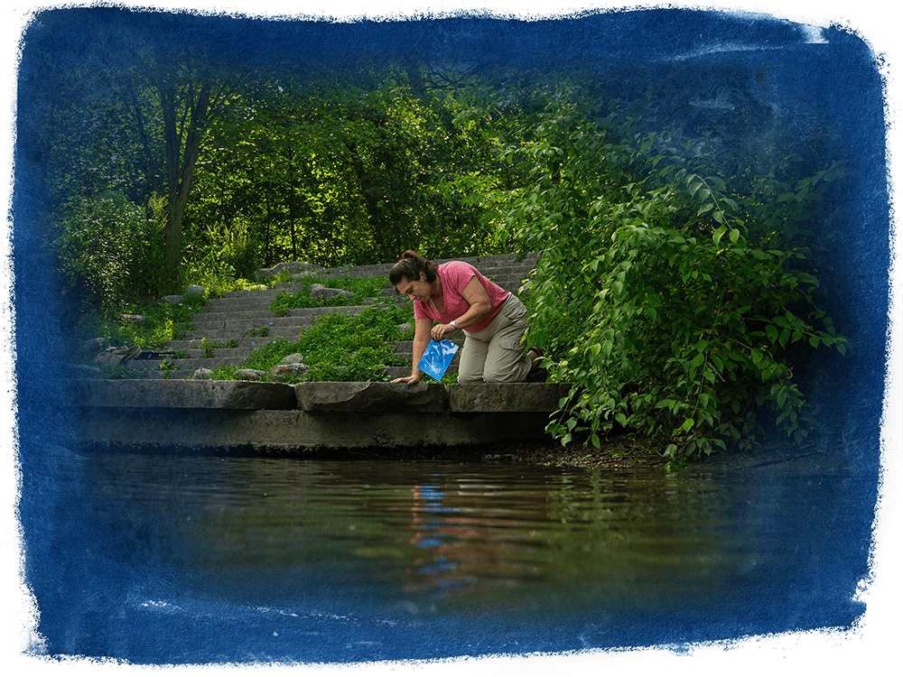 A person in a pink shirt and khaki pants kneels beside the Huron River and pulls a blue piece of paper out of the water. 