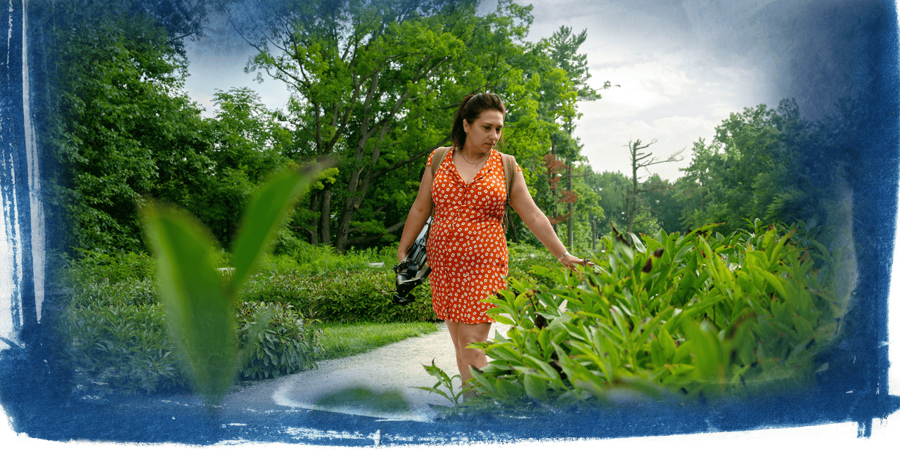 A person in an orange dress walks through an outdoor space with green foliage.