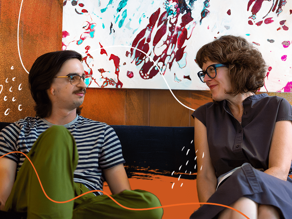A young man wearing glasses and a striped shirt sits next to a woman wearing a gray dress on a couch.