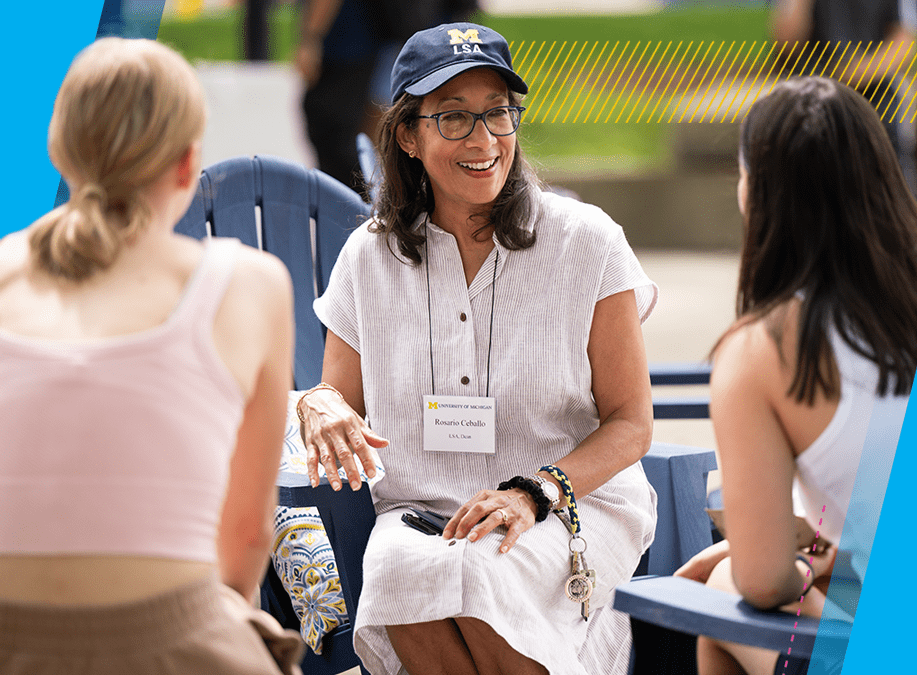 Dean Rosario Ceballo talks with two students during an outdoor event. She is wearing a baseball cap with a Block M and the letters LSA. 