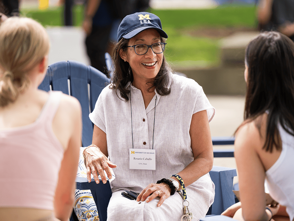 Dean Rosario Ceballo talks with two students during an outdoor event. She is wearing a baseball cap with a Block M and the letters LSA. 