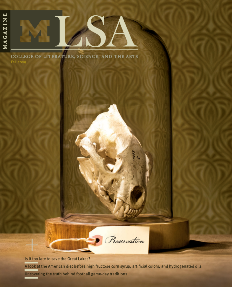 Cover of the Fall 2009 issue of LSA Magazine from the University of Michigan’s College of Literature, Science, and the Arts. The main image features an animal skull, likely that of a mammal, displayed upright under a glass dome on a wooden base. A handwritten label reading “Preservation” is attached to the base. The background shows a patterned, gold-colored wallpaper. Featured article titles at the bottom mention topics such as saving the Great Lakes, the American diet before certain additives, and football game-day traditions. The large “MLSA” logo is at the top, with “MAGAZINE” running vertically along the left edge.