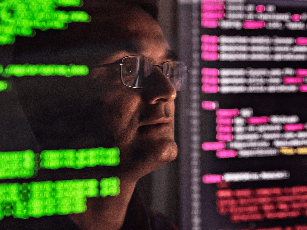 Professor Ambuj Tewari, wearing thin glasses, looks stoically in the distance to the right, angled towards the viewer. The background is different colored code on a computer screen.