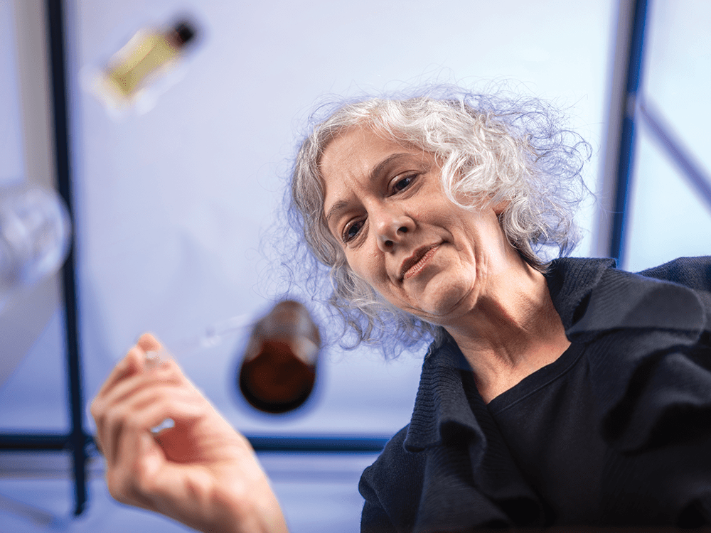 In a photo taken from below, looking up under a clear table, Michelle Kydd takes a liquid out of a small, brown scent bottle using a pipette. Her face looking at the bottle’s opening is in focus.