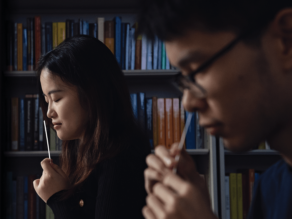 Students Rui Nie and Yikun Han stand in front of a bookcase. They each smell a thin, white smelling strip.