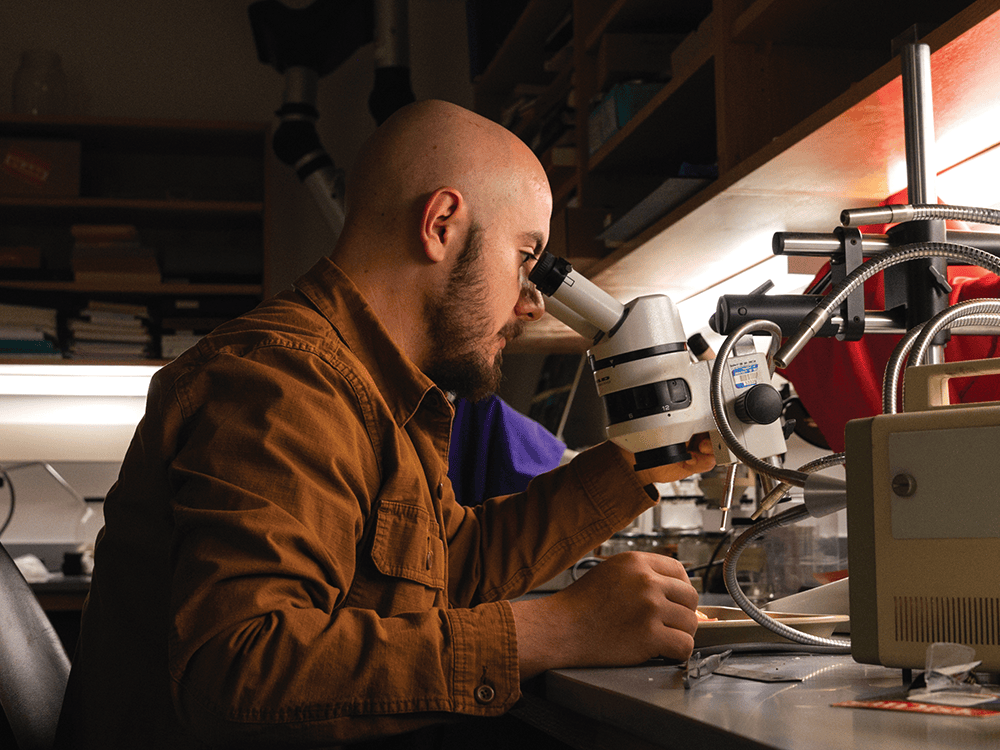 Rodrigo Figueroa is shown in profile, from the waist up, sitting at a gray table and looking into a microscope. Figueroa wears a brown cargo jacket.