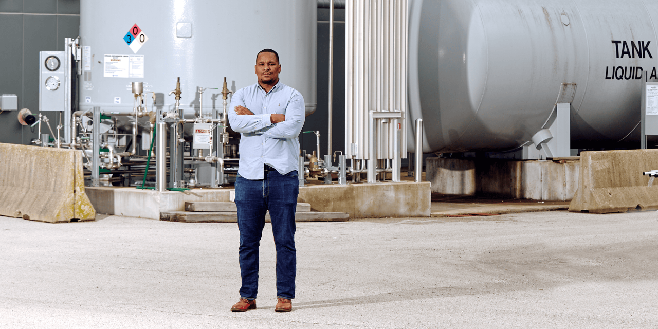 A person with his arms crossed stands in front of machinery in a warehouse space.