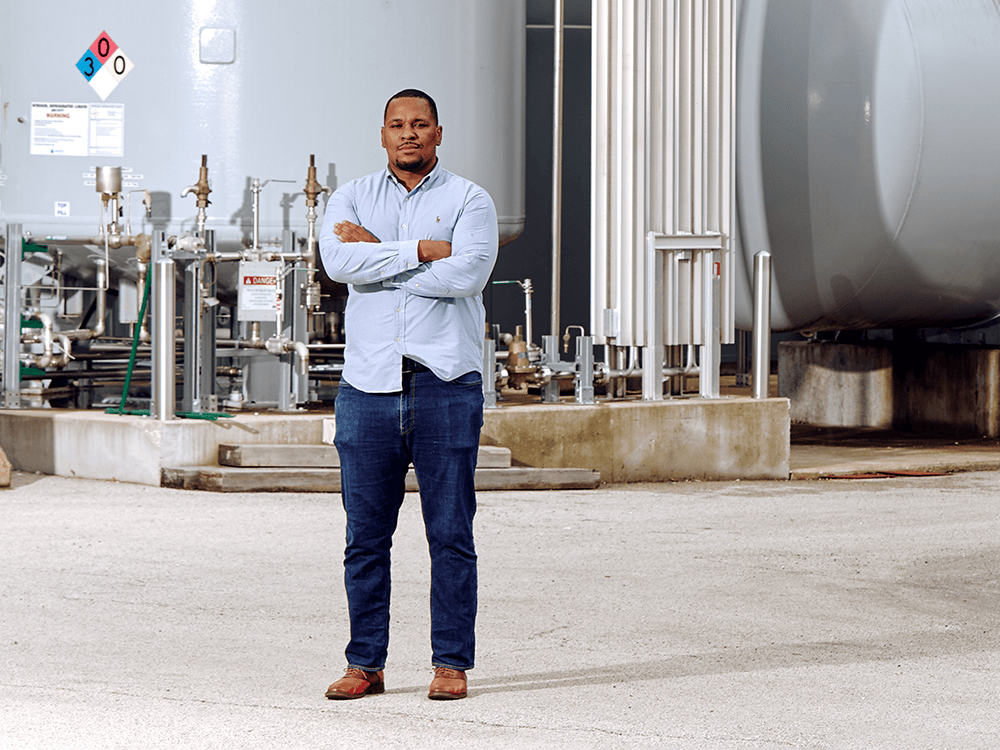 A person with his arms crossed stands in front of machinery in a warehouse space.