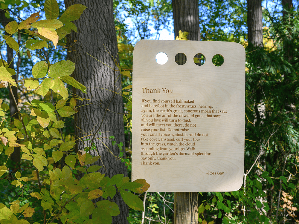 A wooden placard featuring the text of the poem “Thank You,” by Ross Gay, surrounded by trees.