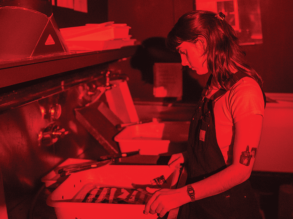 A photograph of a student in profile, inspecting prints. The light is red to emulate a working darkroom. 