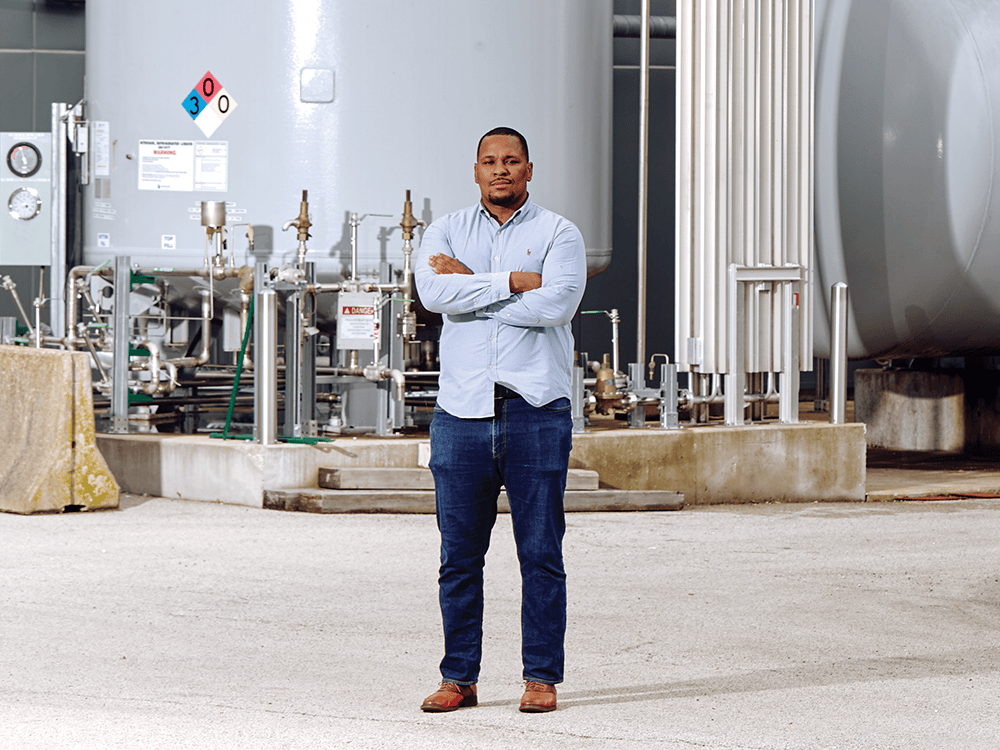 A person with his arms crossed stands in front of machinery in a warehouse space.