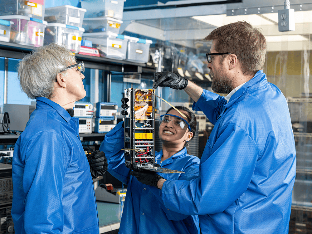 Three researchers wearing blue lab coats and black gloves examine a one-foot-tall boxy satellite.