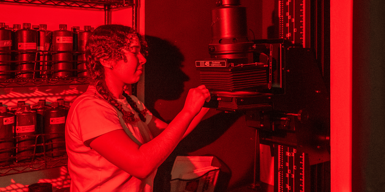 A photograph of a student in profile, working with a photo enlarger carrier. The light is red to emulate a working darkroom.