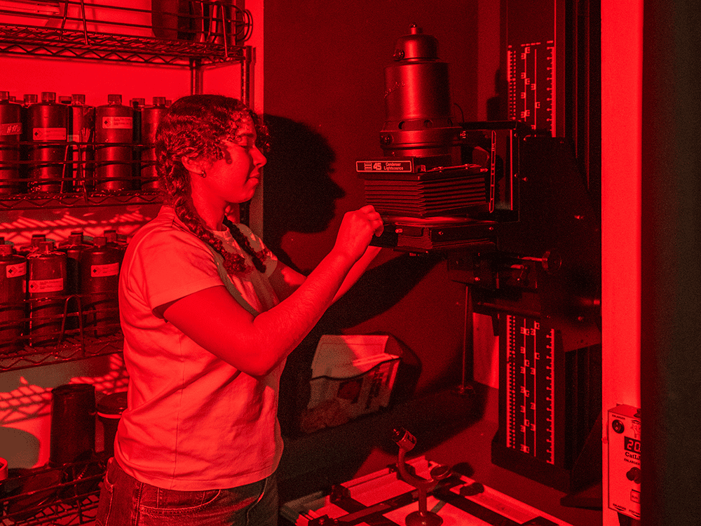 A photograph of a student in profile, working with a photo enlarger carrier. The light is red to emulate a working darkroom.