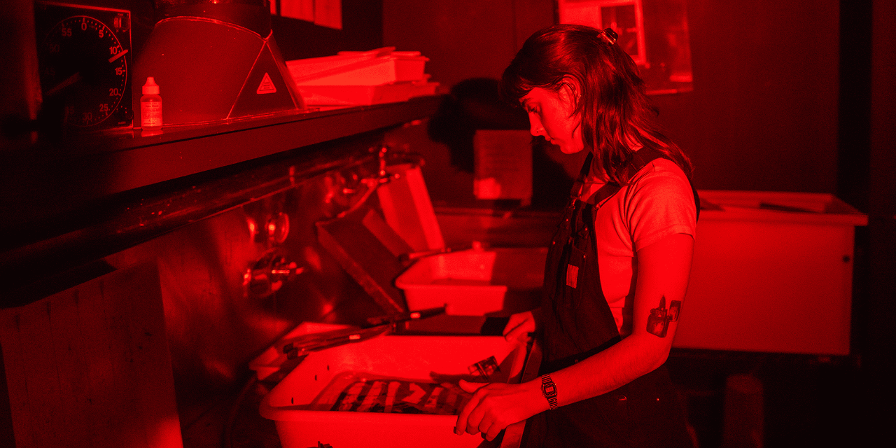 A photograph of a student in profile, inspecting prints. The light is red to emulate a working darkroom.  