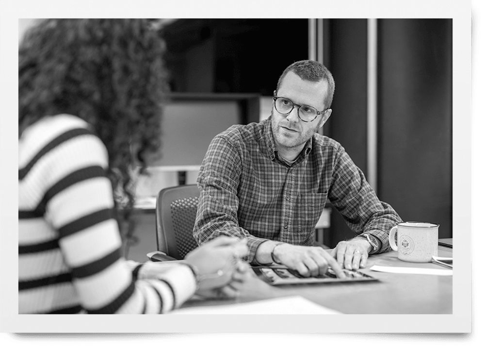 A black and white photograph of a faculty member meeting with a student. They are seated at a large table, examining a print.