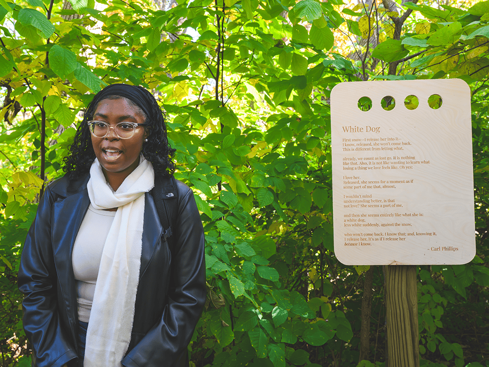 A photograph of a student in glasses beside a wooden placard on which Carl Phillips’s poem “White Dog” is inscribed. 