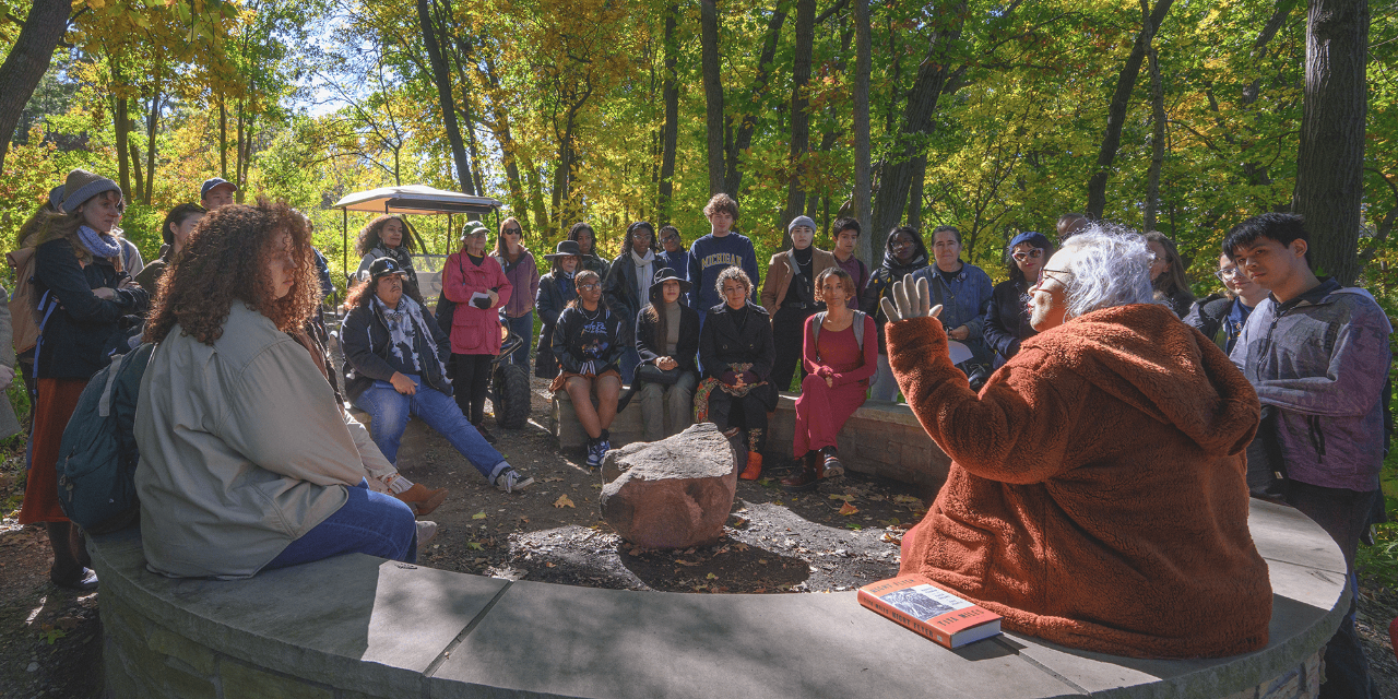 A photograph of a large group of people seated in a circle in a wooded area. A person in a rust-colored jacket tells a story. 