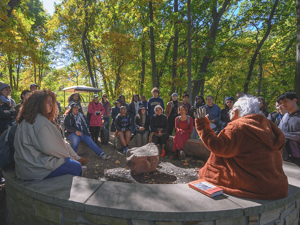 A photograph of a large group of people seated in a circle in a wooded area. A person in a rust-colored jacket tells a story. 